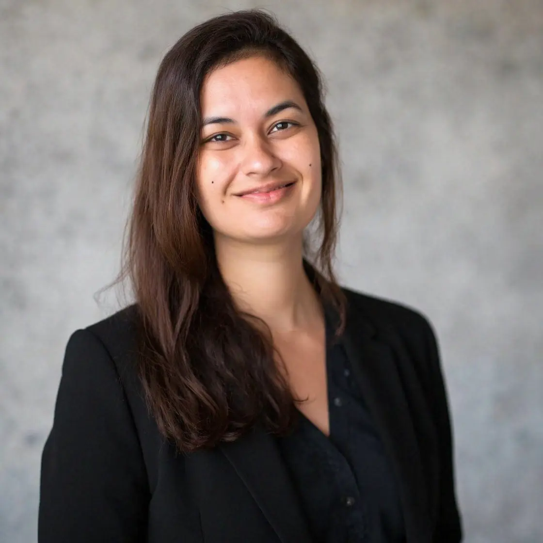 A professional portrait of Soureya wearing a dark blazer in front of a modern office